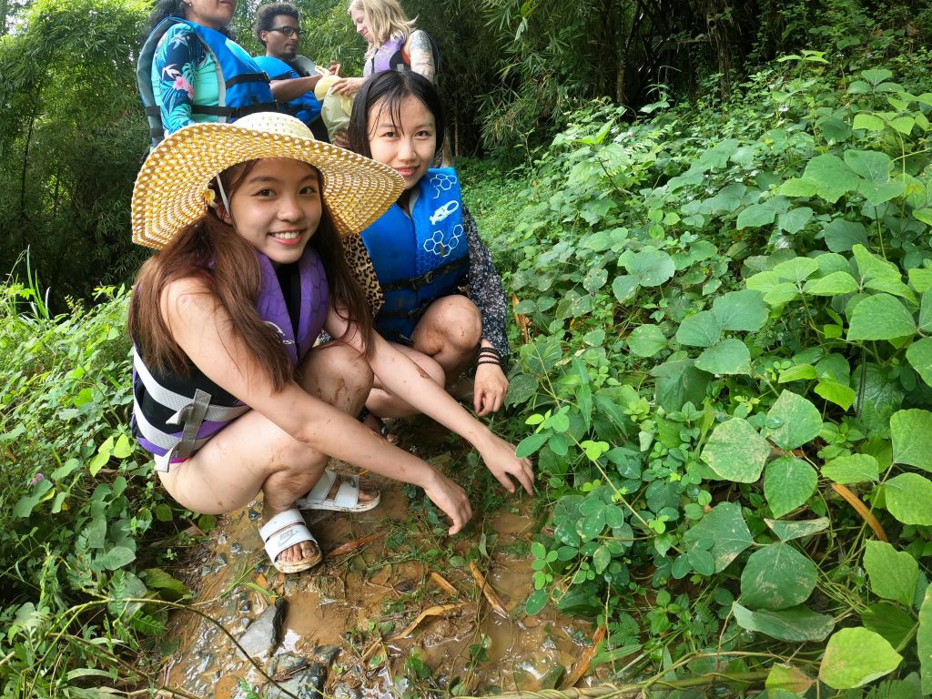 구명조끼와 밀짚모자를 쓴 두 여성이 녹지에 웅크린 채 미소를 짓고 있습니다. 구명조끼를 입은 다른 사람들이 그 뒤에 보입니다.
