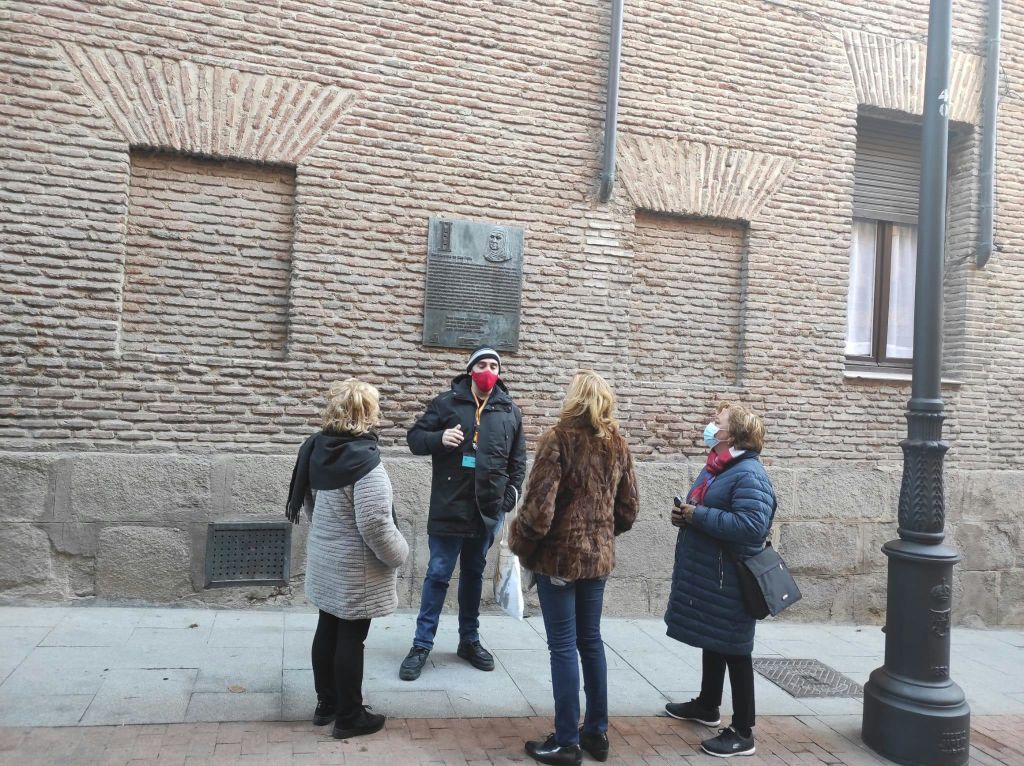 명판이 걸린 벽돌 벽 앞에서 네 명이 인도에 서서 대화를 나누고 있습니다. 두 개는 마스크를 쓰고, 한 개는 커피를 담습니다.