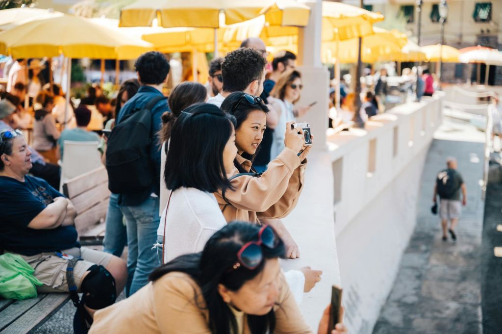 사람들은 노란 우산 아래 야외에 모입니다. 다른 사람들이 근처에 앉거나 서 있는 동안 여성은 카메라에 초점을 맞춥니다.