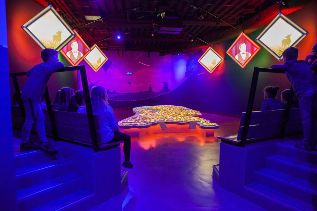 벽에 조명 패널이 있고 바닥에 커다란 빛나는 모양이 있는 어두운 방에서 사람들은 층층이 쌓인 좌석에 앉아 있습니다.