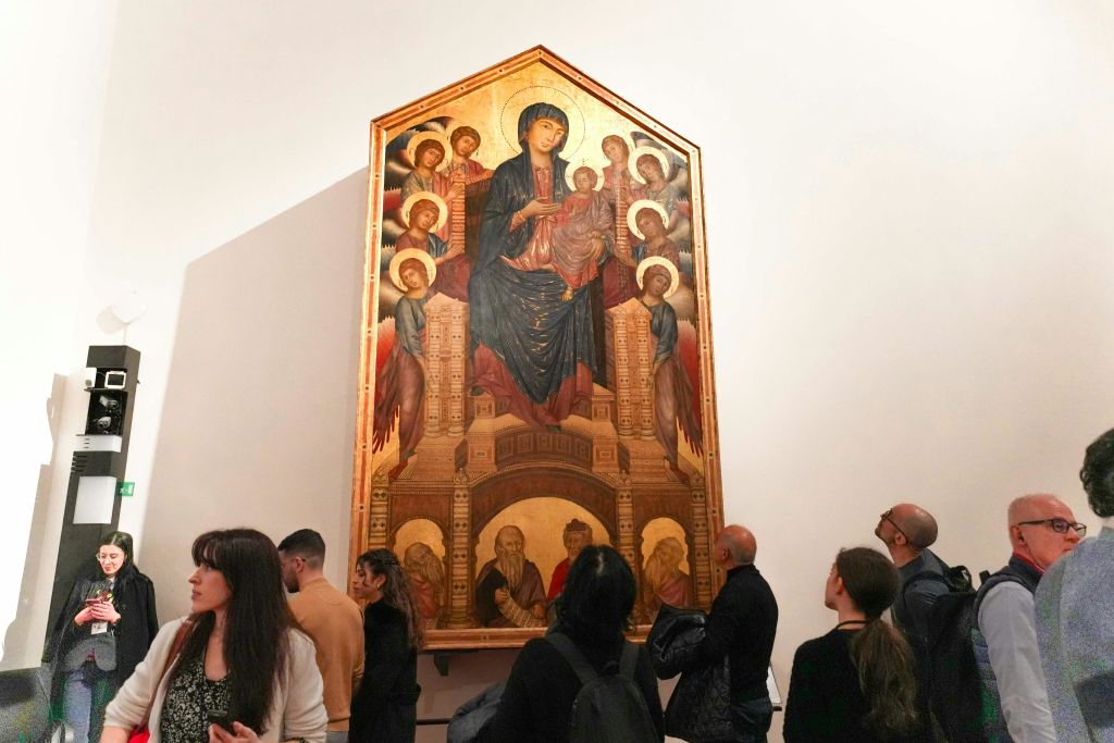 여러 인물과 황금색 악센트가 있는 종교적 장면을 묘사한 박물관에서 중세 시대의 대형 그림을 보는 사람들.