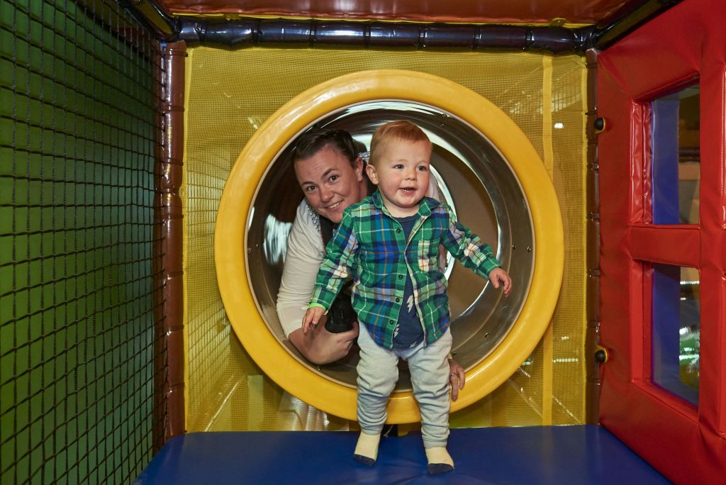 격자무늬 셔츠를 입은 웃는 유아가 실내 놀이터의 노란색 놀이 터널에서 한 여성 앞에 서 있습니다.