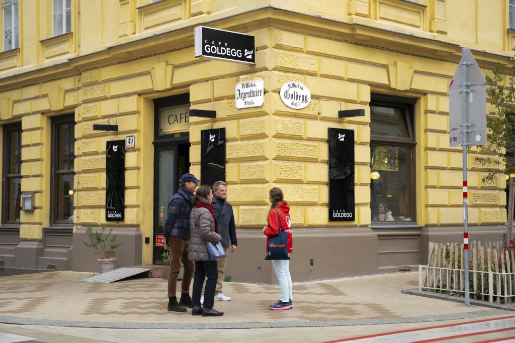 검은색 간판과 장식 요소가 있는 노란색 건물인 카페 골데그 외부에서 네 명이 서서 이야기를 나누고 있습니다.