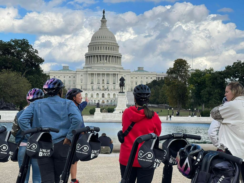 부분적으로 흐린 하늘 아래 미국 국회의사당 건물을 마주보고 있는 헬멧과 스쿠터를 착용한 사람들.