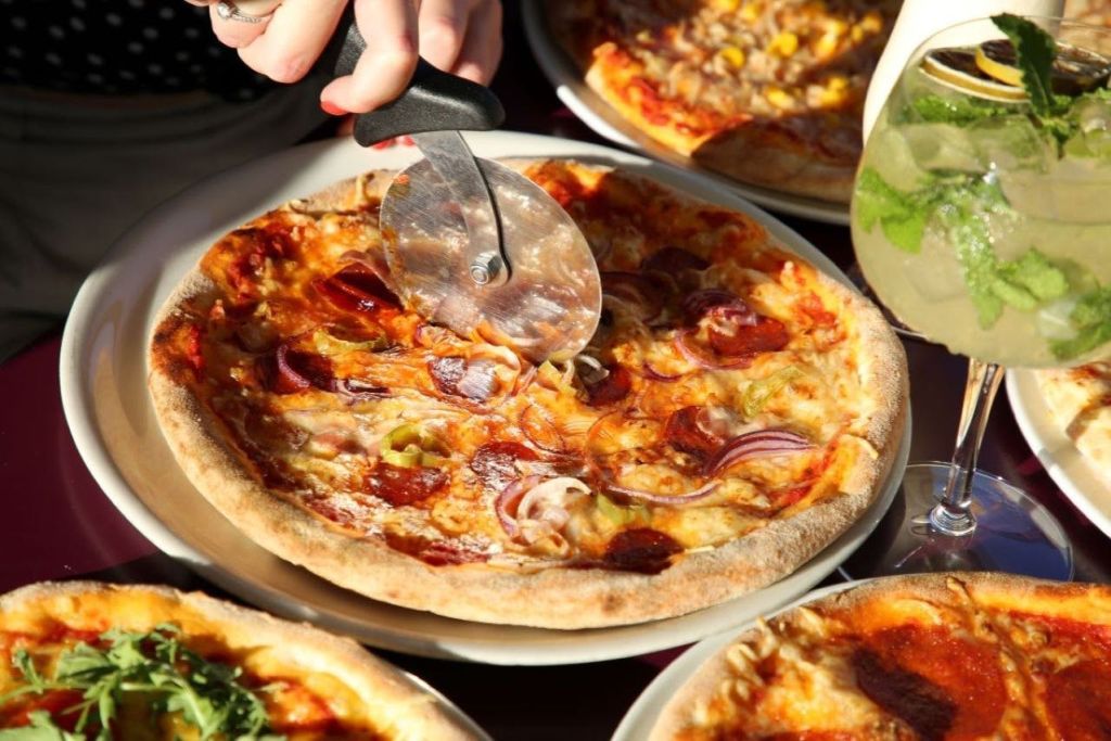 한 사람이 피자 커터를 사용하여 페퍼로니, 양파, 청양고추를 얹은 피자를 썰고 있습니다. 근처에 다른 피자와 음료가 있습니다.