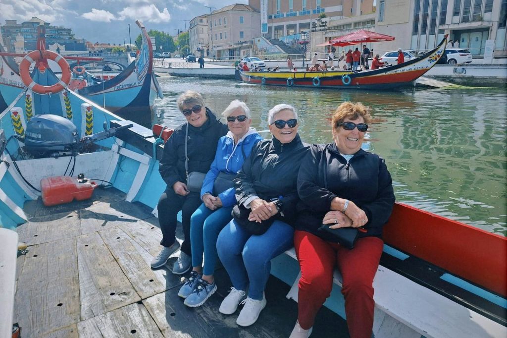 운하가 정박한 보트에 선글라스와 재킷을 착용한 여성 4명이 앉아 있으며, 화려한 건물과 보트가 배경으로 펼쳐집니다.