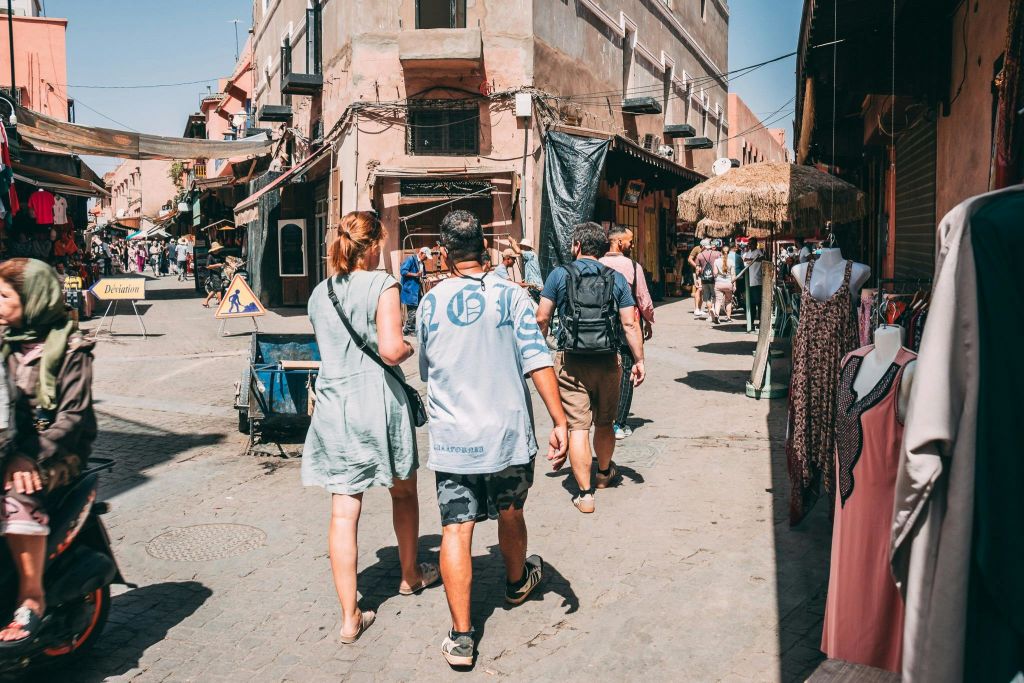 화창한 하늘 아래 소박한 건물, 상점, 다양한 상품이 진열된 번화한 시장 거리를 걷는 사람들.