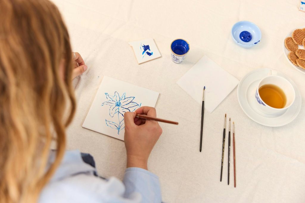 붓, 작은 페인트 컵, 또 다른 페인트 타일로 둘러싸인 타일에 파란색 꽃을 그리는 사람.