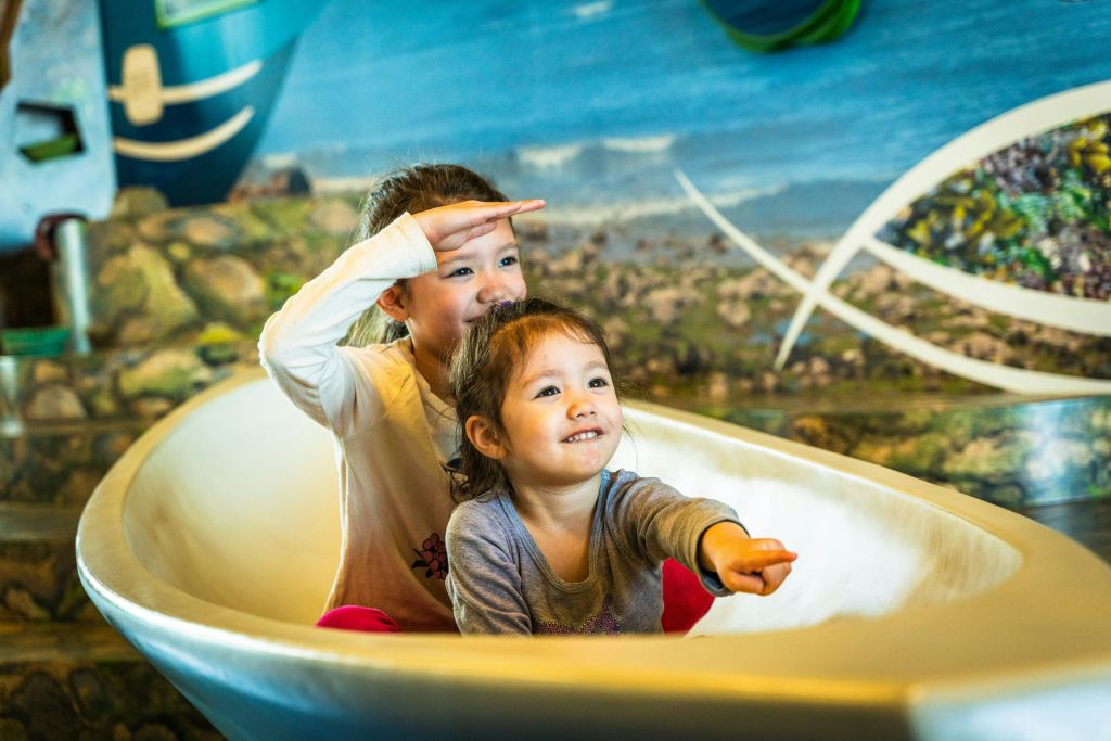 두 명의 어린이가 하얀 보트 모양의 구조물에 앉아 다채로운 벽화를 배경으로 항해하는 척하고 있습니다.