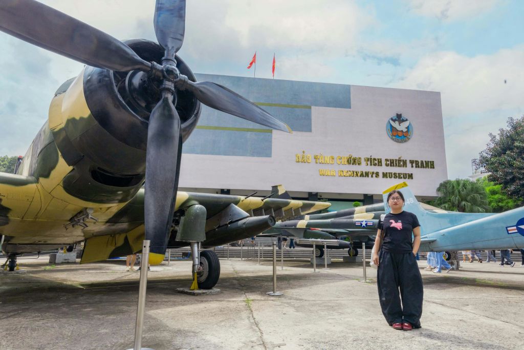 박물관 건물과 깃발이 보이는 베트남 전쟁 잔존물 박물관에서 한 사람이 항공기 앞에 서 있습니다.