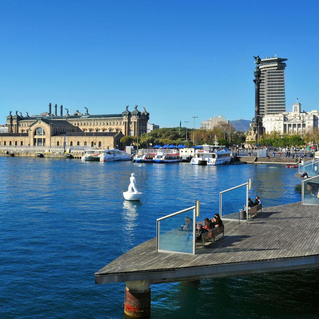 맑고 푸른 하늘 아래 보트와 유서 깊은 건물이 내려다보이는 수변 데크에 사람들이 앉아 있습니다.