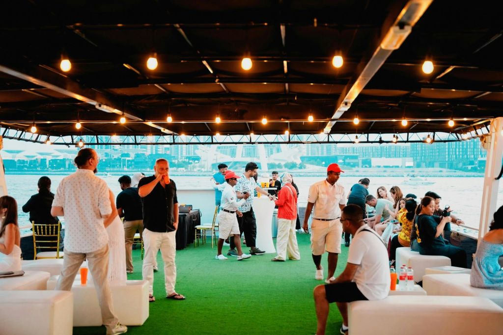 인공 잔디가 깔린 보트 데크에서 사교 활동을 즐기는 사람들, 머리 위에는 현악기 조명이 있고, 수변 도시 풍경을 배경으로 활동하는 사람들.