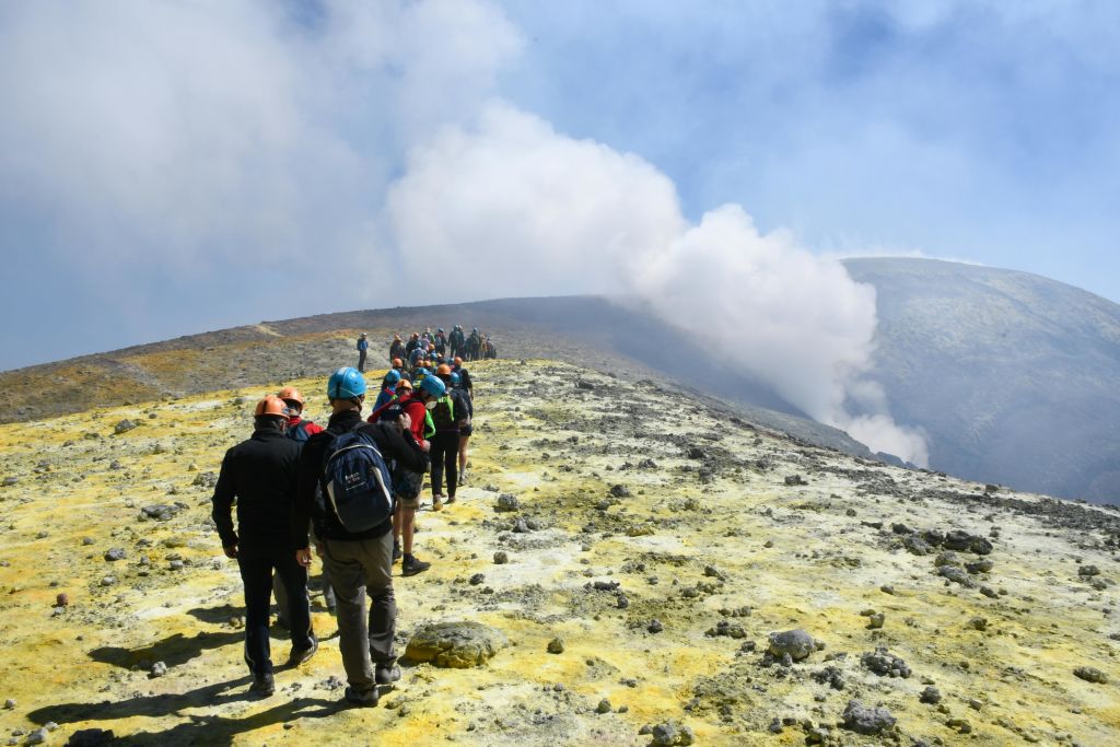 한 무리의 사람들이 헬멧과 배낭을 착용하고 바위투성이의 황색 지형을 따라 하이킹을 하고 있으며, 연기가 솟아오르고 있습니다.