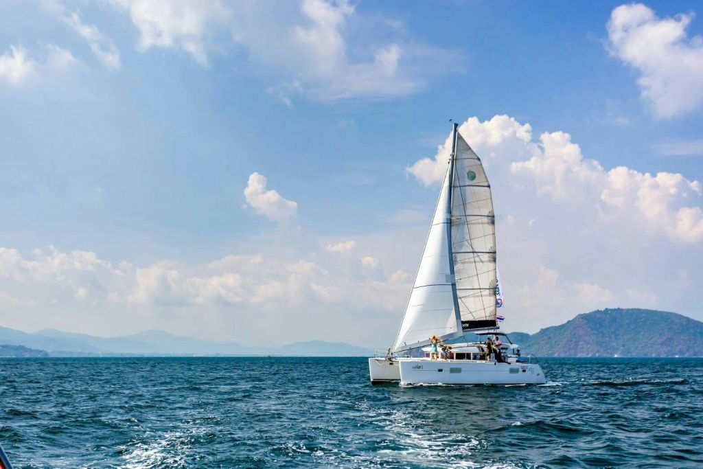 먼 산을 배경으로 부분적으로 흐린 하늘 아래 잔잔한 바다에서 돛을 올린 하얀 요트.