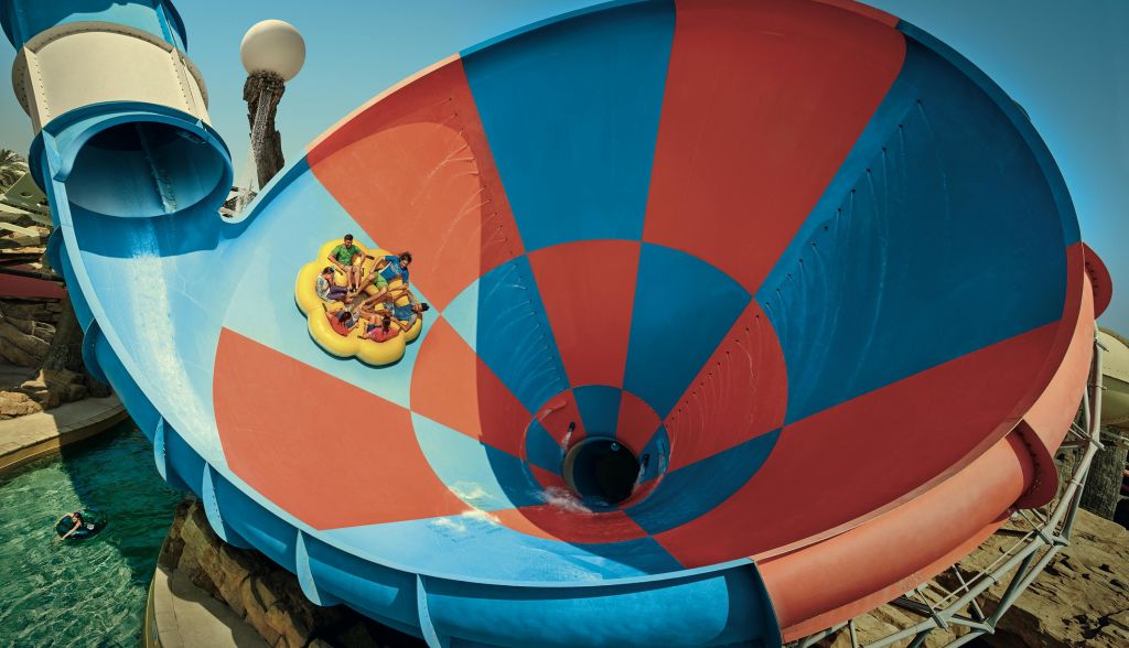 야외 워터파크의 깔때기 모양의 크고 화려한 워터 슬라이드에 노란색 뗏목을 타는 사람들.