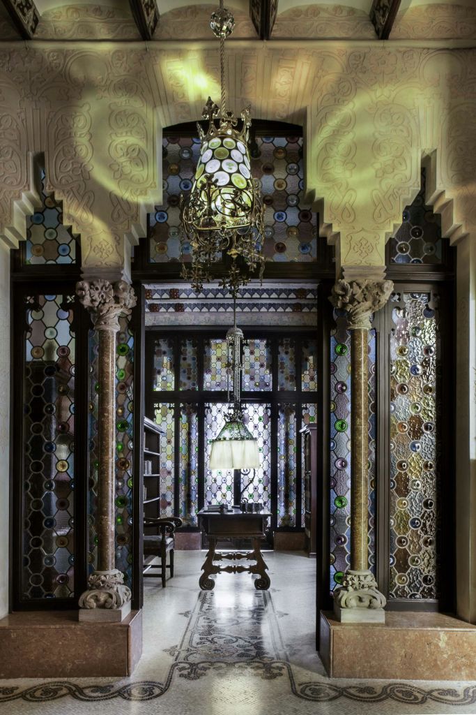 화려한 스테인드글라스, 정교한 몰딩, 기둥, 샹들리에, 중앙에 램프가 있는 나무 책상이 있는 화려한 객실입니다.