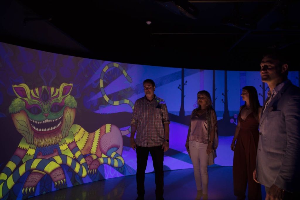 벽에 뱀과 정글 배경의 화려한 애니메이션 투영으로 조명이 켜진 어두운 방에 어른 네 명이 서 있습니다.