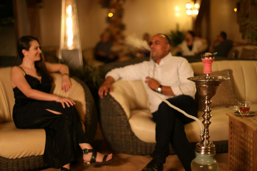 따뜻한 조명으로 꾸며진 방에서 한 여성과 남성이 고리버들 안락의자에 앉아 대화를 나누고 있습니다. 배경에 스탠딩 히터가 보입니다.