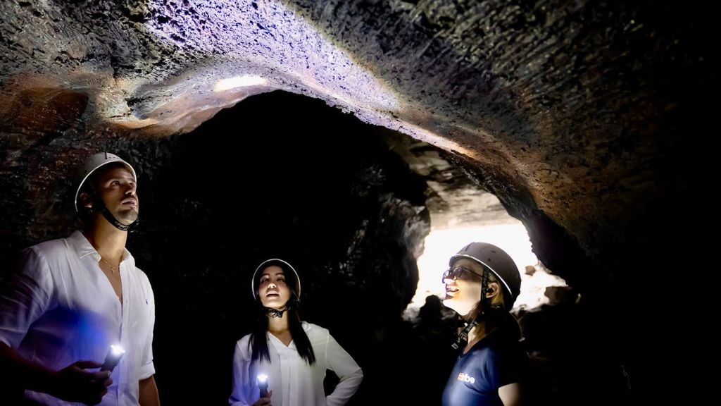 헬멧과 전조등을 착용한 세 사람이 조명이 어두운 동굴을 탐험하며 손전등으로 동굴 천장을 비춥니다.