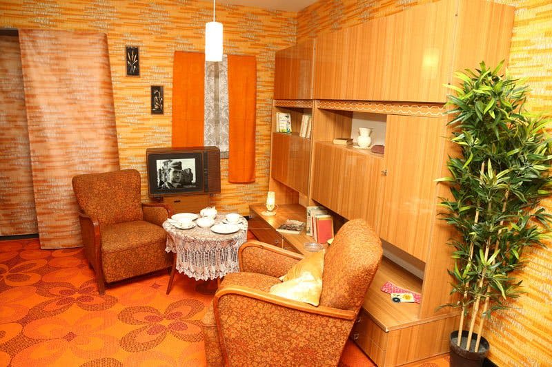 주황색 가구와 인테리어가 돋보이는 복고풍 거실, 흑백 이미지를 보여주는 TV, 차 세트가 놓인 작은 테이블, 나무 수납장이 있습니다.