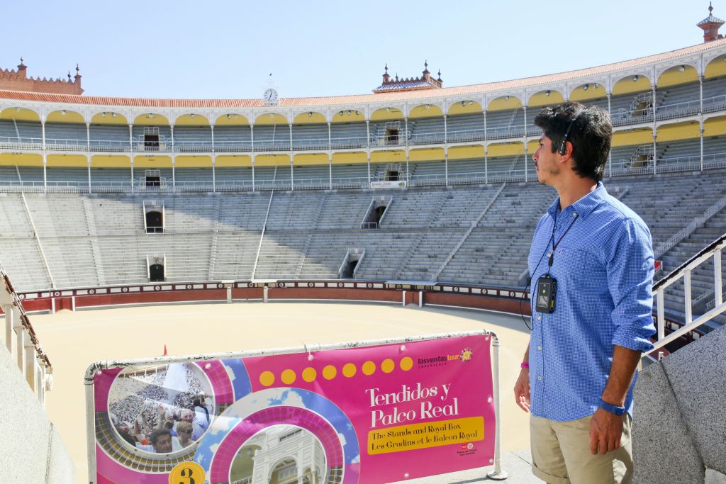 오디오 가이드를 든 남성이 빈 황소 고리에 앉아 좌석을 바라보고 있습니다. 분홍색 간판에는 "Tendidos y Palco Real" 이라고 쓰여 있습니다.
