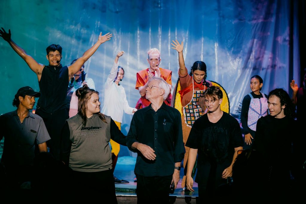 화려한 의상과 캐주얼한 복장을 한 사람들이 무대 위에 서 있으며, 일부는 파란색 커튼을 배경으로 손을 들어 올립니다.