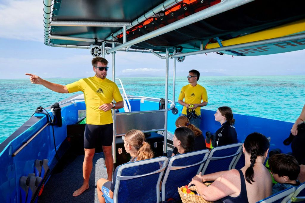 노란색 셔츠를 입은 두 명의 강사가 맑고 청록색 바다 근처의 보트에 앉은 네 명의 사람들에게 브리핑을 합니다.