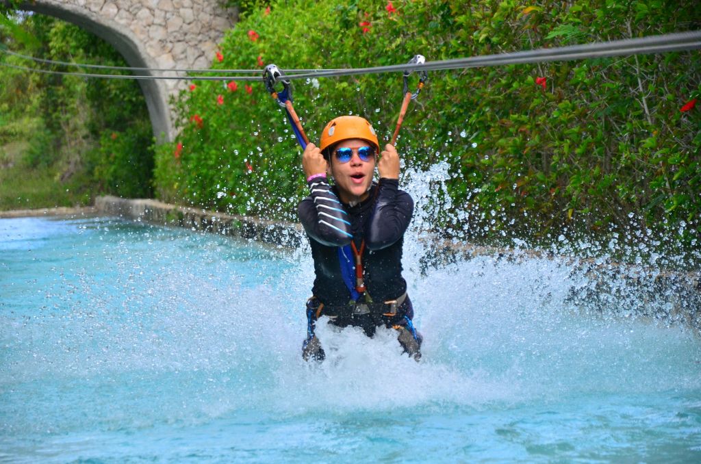 짚라인을 탄 사람이 헬멧과 선글라스를 쓰고 수영장에 뛰어들고 있으며, 녹지와 석조 아치를 배경으로 하고 있습니다.
