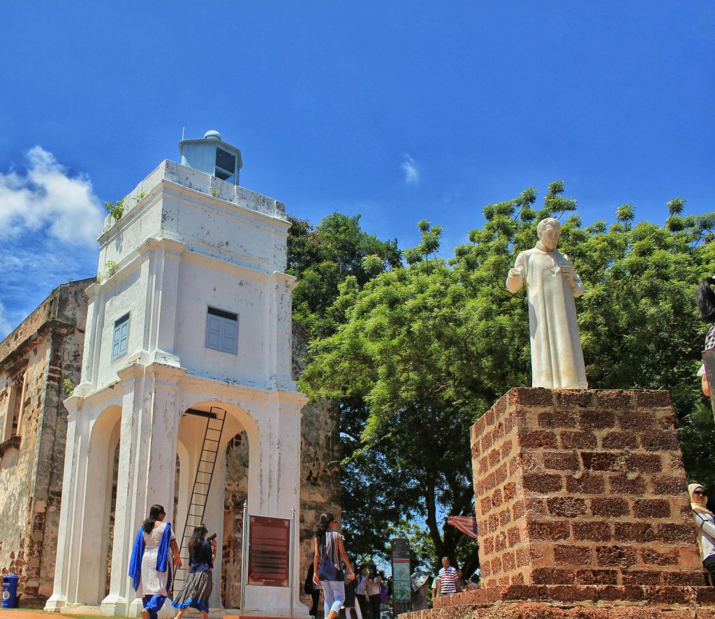 나무와 푸른 하늘을 배경으로 높은 흰색 종탑과 석조 받침대 위의 동상 근처를 걷는 사람들.