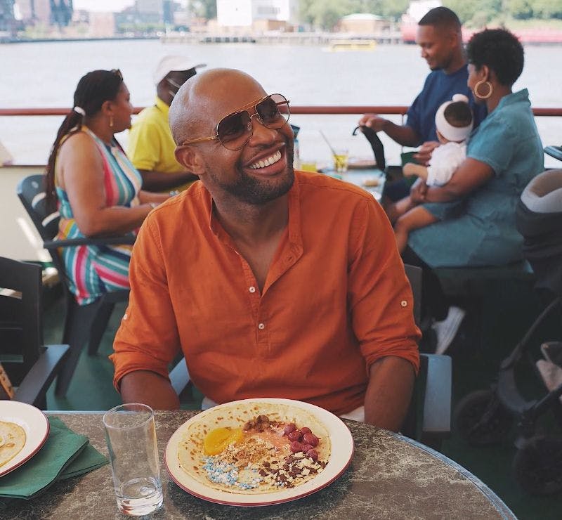 주황색 셔츠를 입은 웃는 사람이 부분적으로 식사를 한 테이블에 앉아 있습니다. 다른 사람들이 둘러앉아 아기를 안고 있습니다.