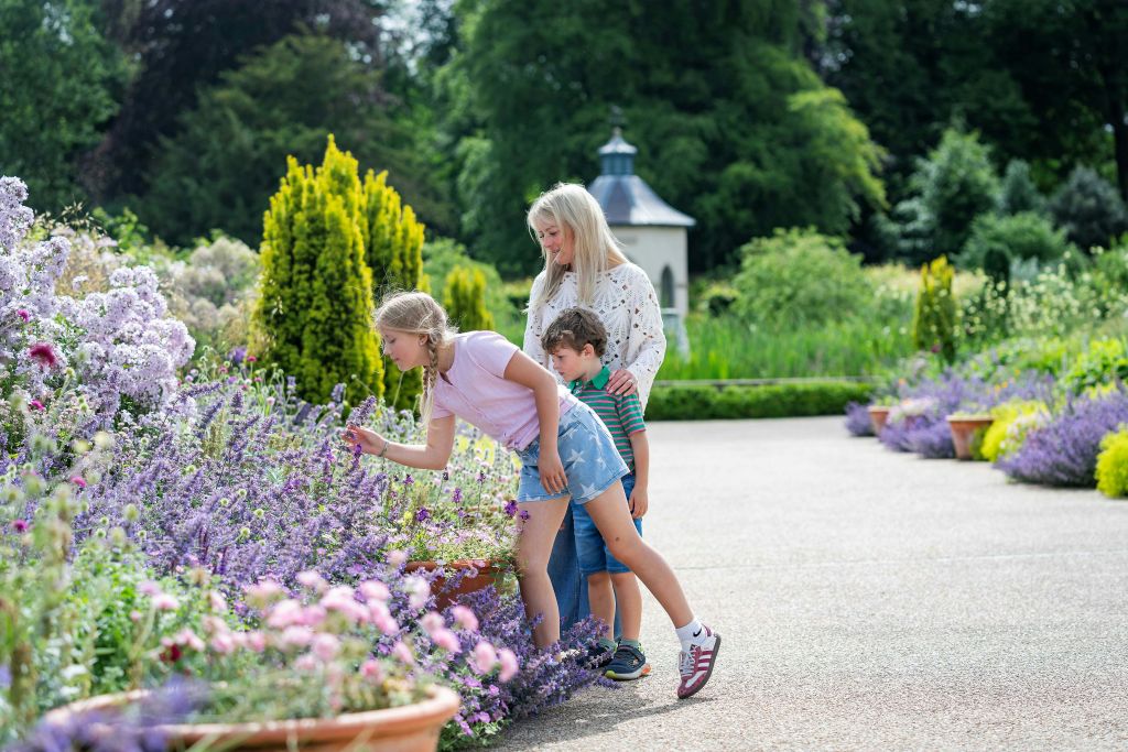 푸르름이 우거진 정원과 작고 하얀 정자를 배경으로 한 여성과 두 명의 어린이가 꽃을 살펴보고 있습니다.