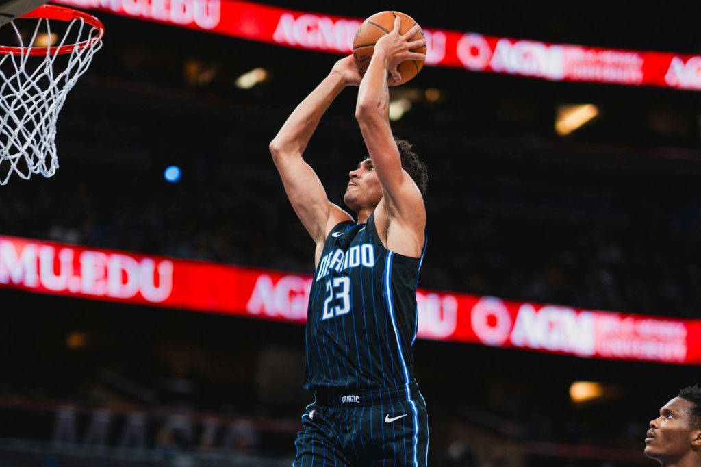 파란색 "올랜도" 유니폼을 입은 농구 선수가 빨간색 LED 디스플레이를 배경으로 경기장에서 공을 쏘기 위해 점프합니다.