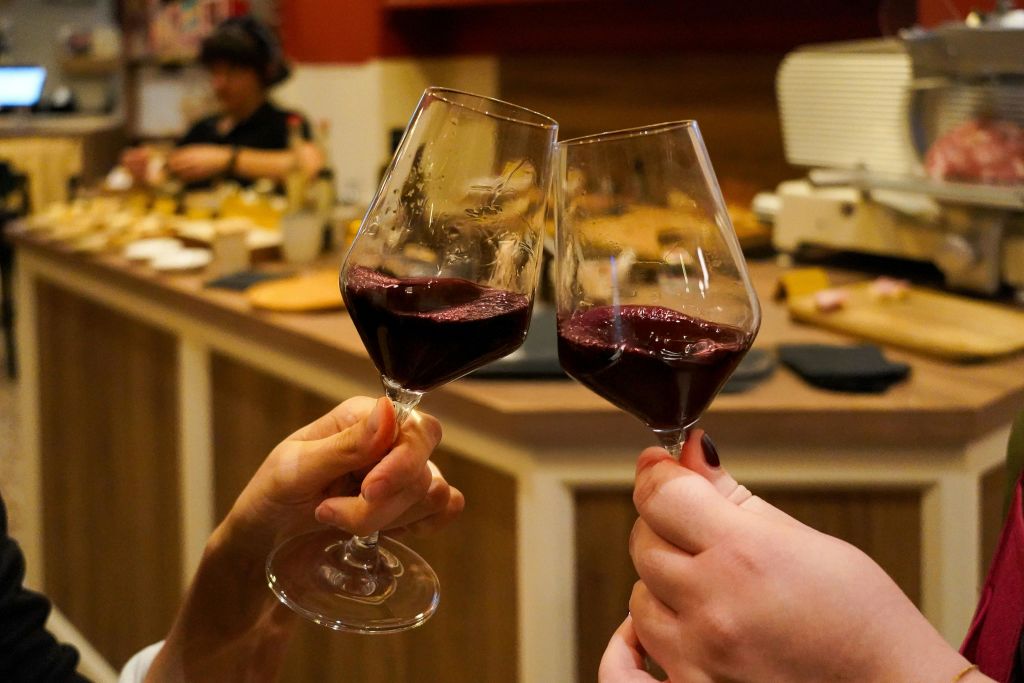 아늑한 실내 분위기에서 레드 와인 잔을 들고 토스트하는 두 손, 사람과 음식을 배경으로 흐릿한 분위기를 자아냅니다.