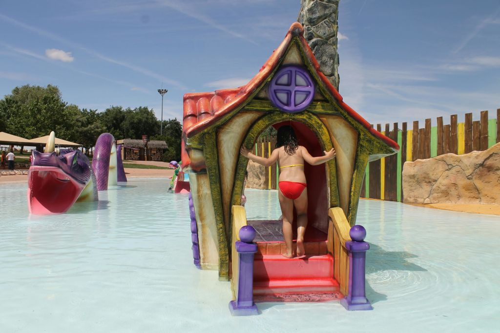 얕은 물놀이 공간의 작고 다채로운 플레이하우스에 빨간 수영복을 입은 어린이가 다른 어린이들과 함께 배경으로 입장합니다.