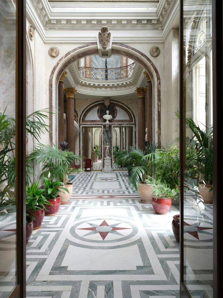 대리석 기둥, 중앙에 동상, 무늬 바닥을 따라 화분이 있는 화려한 복도.