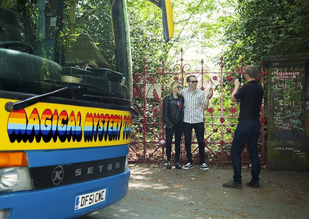 그래피티로 덮인 붉은 대문 옆의 세 사람. 한 남자와 한 여자가 포즈를 취하고 다른 남자가 사진을 찍습니다. 근처에 파란색 버스가 있습니다.