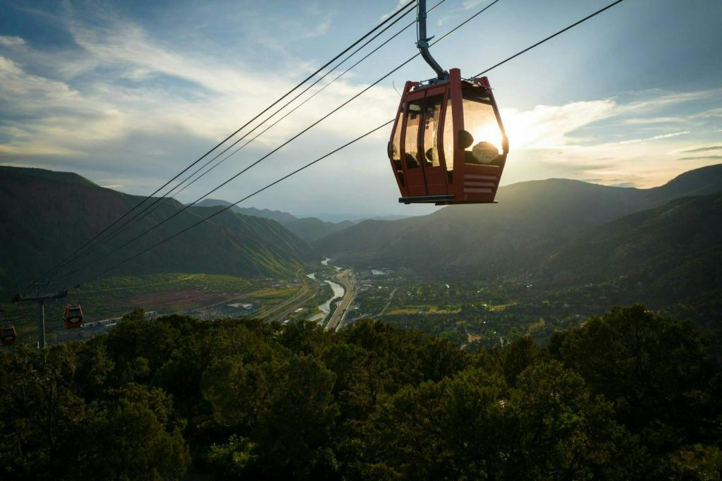 부분적으로 흐린 하늘 아래 산과 구불구불한 강이 있는 울창한 계곡 위에 빨간색 케이블카가 매달려 있습니다.