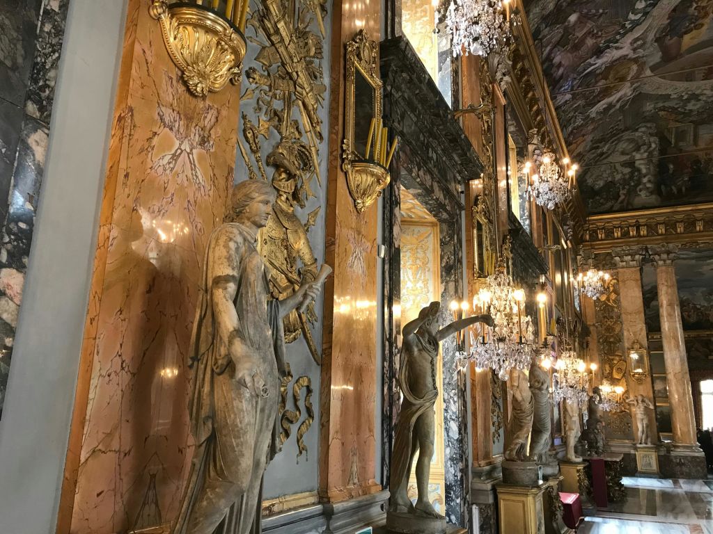 대리석 기둥, 화려한 조각상, 크리스탈 샹들리에, 벽과 천장에 금색 디테일이 있는 고급스러운 홀.