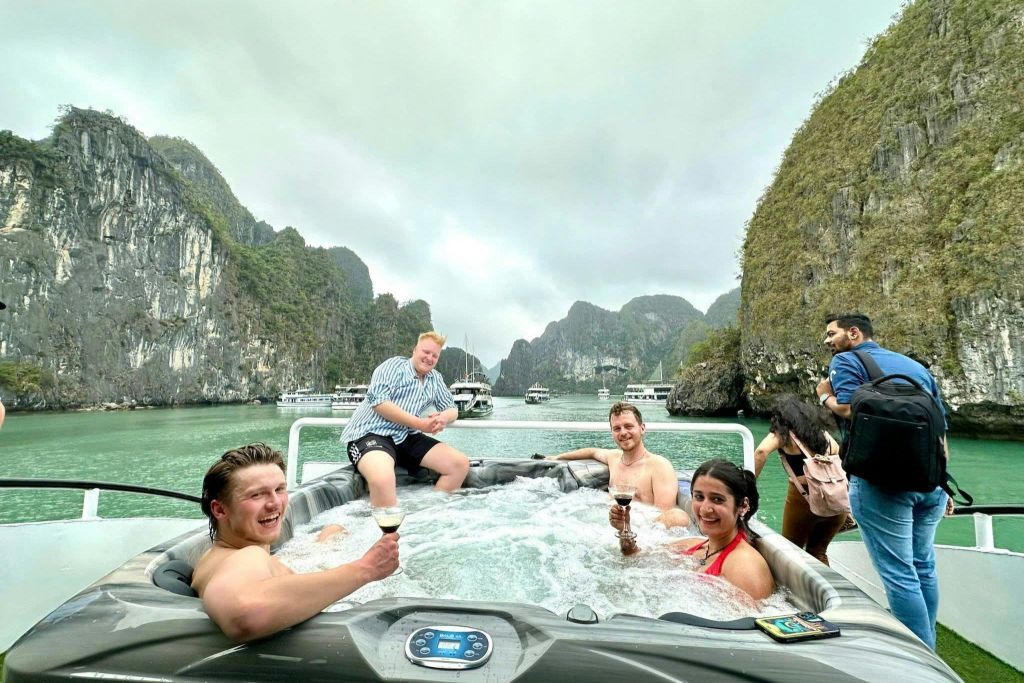 물과 바위 절벽으로 둘러싸인 보트에서 자쿠지에서 휴식을 취하는 수영복을 입은 네 명의 사람들. 그들은 음료를 들고 미소를 짓고 있습니다.