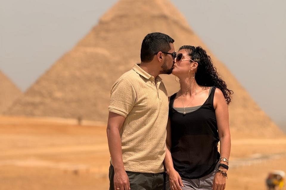화창한 날 피라미드 앞에서 키스하는 커플. 남자는 베이지색 셔츠를 입고, 여자는 검은색 상의와 선글라스를 착용합니다.