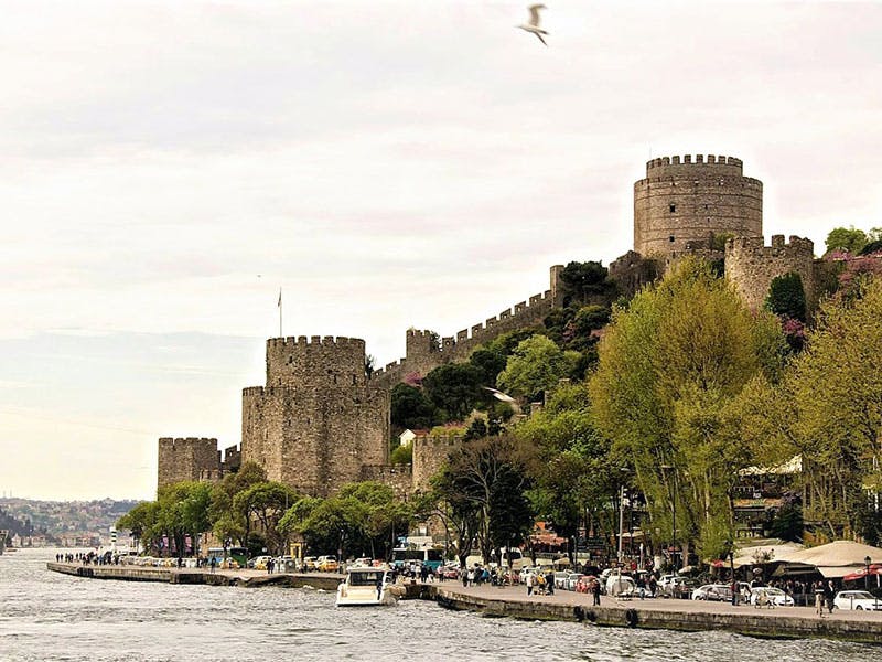 언덕에 원통형 타워가 있는 석조 요새로, 나무로 둘러싸여 있으며 물가를 따라 사람과 보트가 있습니다.