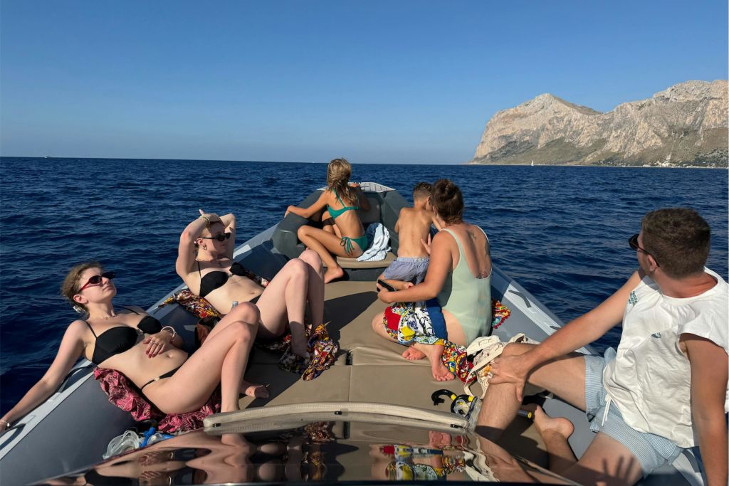 탁 트인 바다에서 보트에서 일광욕을 즐기고 휴식을 취하는 사람들. 맑은 하늘 아래 배경으로 바위 해안선이 보입니다.