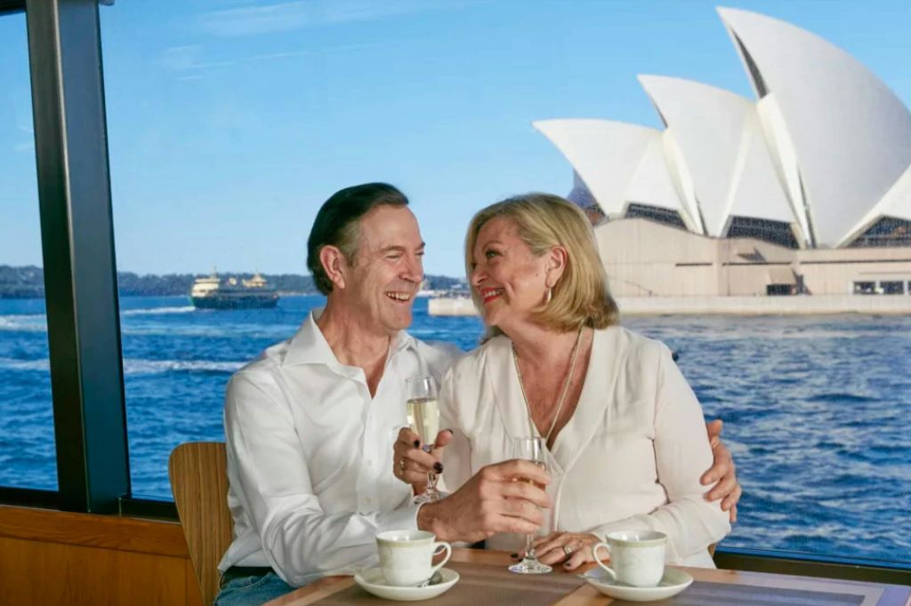 커피잔이 놓인 테이블에서 샴페인잔을 들고 미소를 짓고 있는 부부. 시드니 오페라하우스가 배경으로 보입니다.