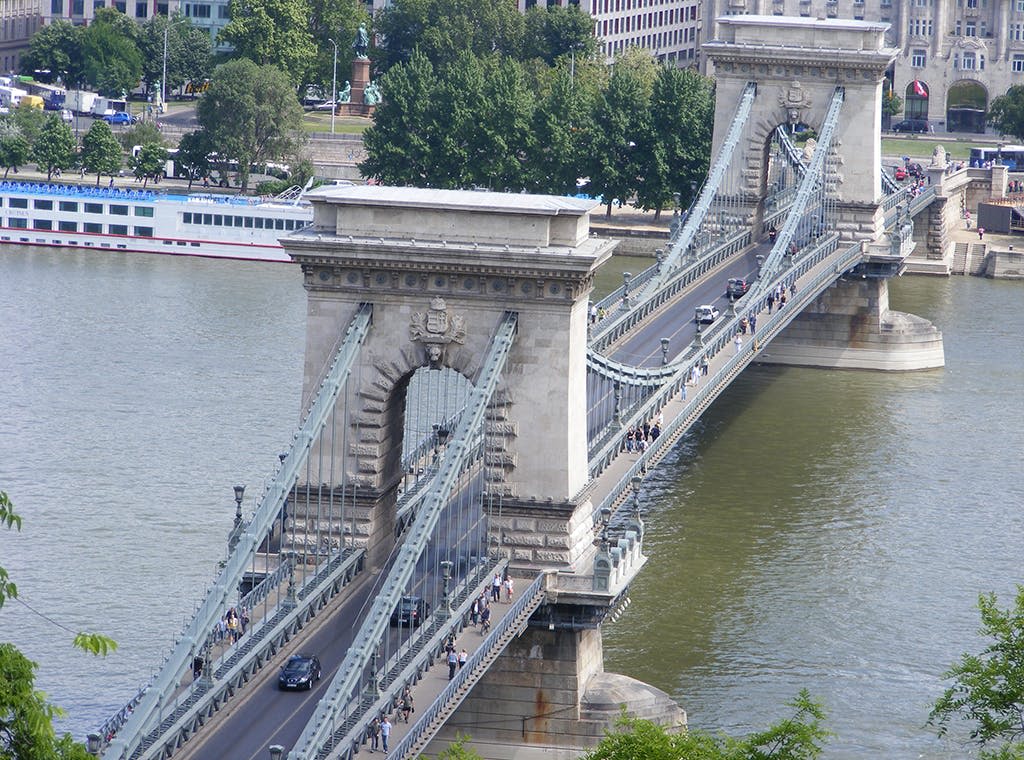석탑이 있는 현수교가 강에 걸쳐 있으며, 차량과 보행자가 건넌다. 나무와 건물이 배경으로 보입니다.