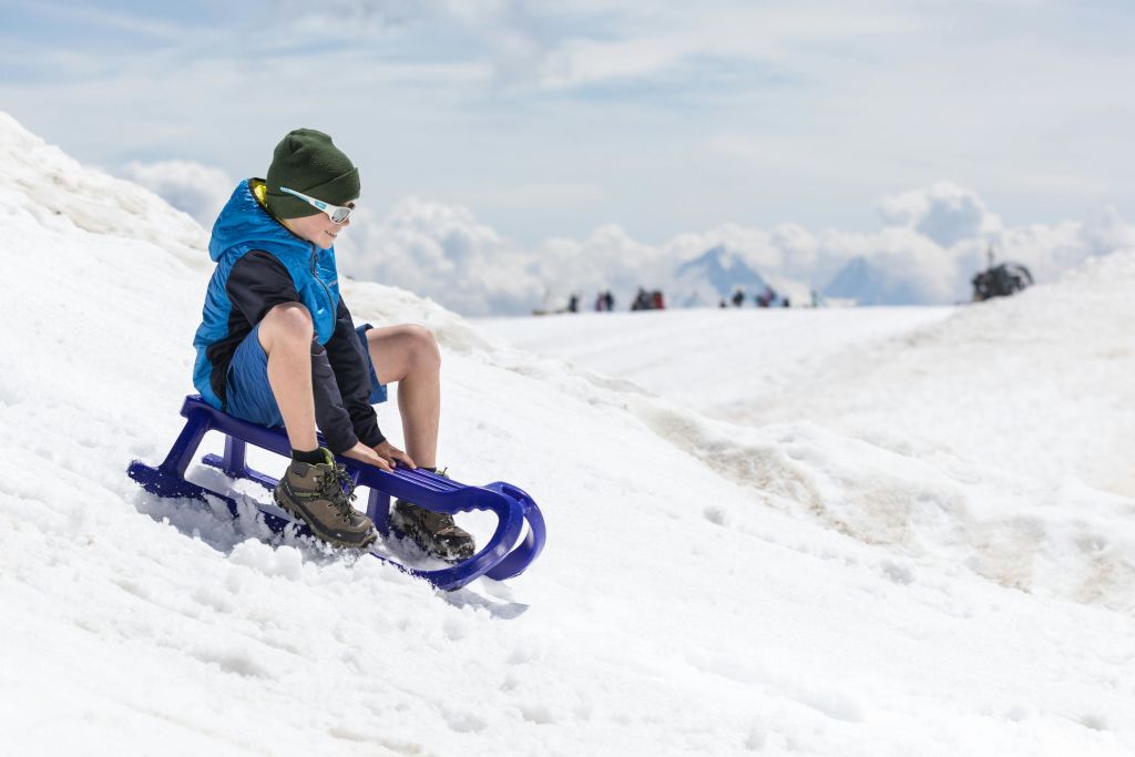 겨울옷을 입은 어린이가 눈 덮인 경사면 꼭대기에서 보라색 썰매를 타고 산과 부분적으로 흐린 하늘을 배경으로 앉아 있습니다.
