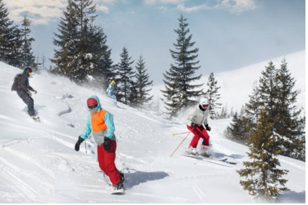형형색색의 장비를 착용한 세 명의 스키어가 부분적으로 흐린 하늘 아래 눈 덮인 나무가 늘어선 비탈을 따라 내려갑니다.