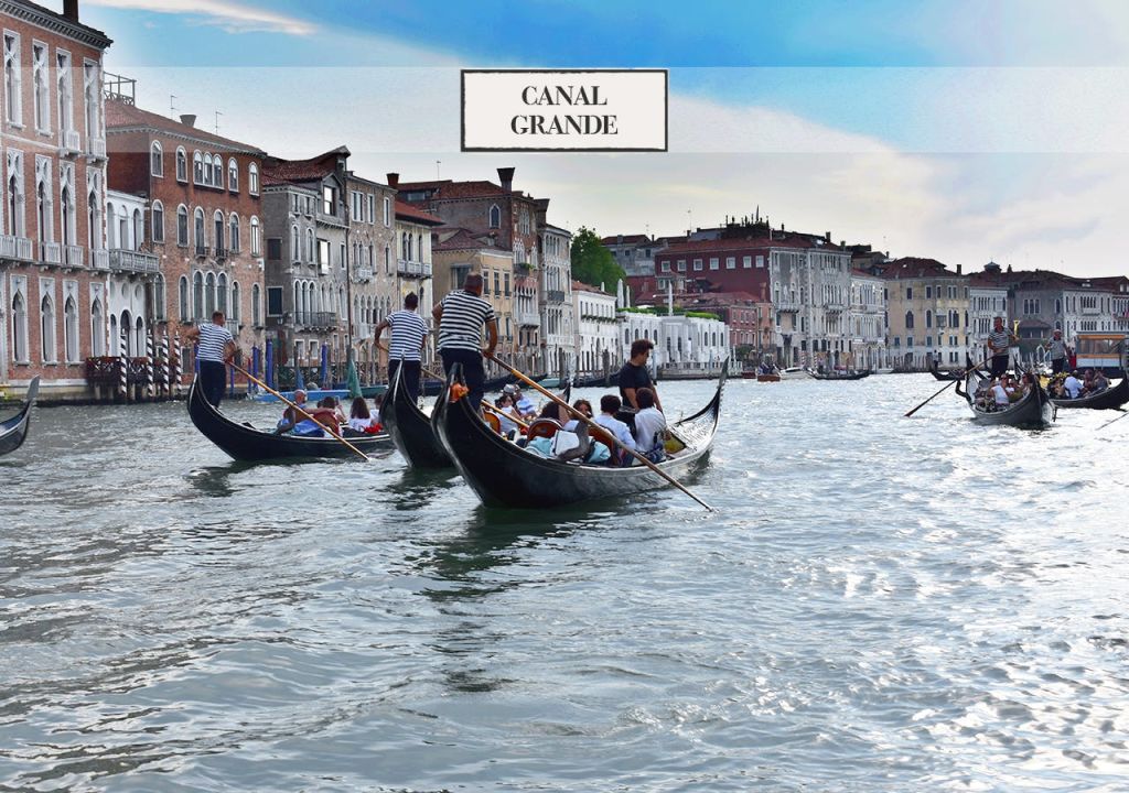 푸른 하늘 아래 역사적인 건물이 늘어선 운하를 따라 승객과 함께 곤돌라가 떠 있습니다. 간판에는 "Canal Grande" 라고 쓰여 있습니다.