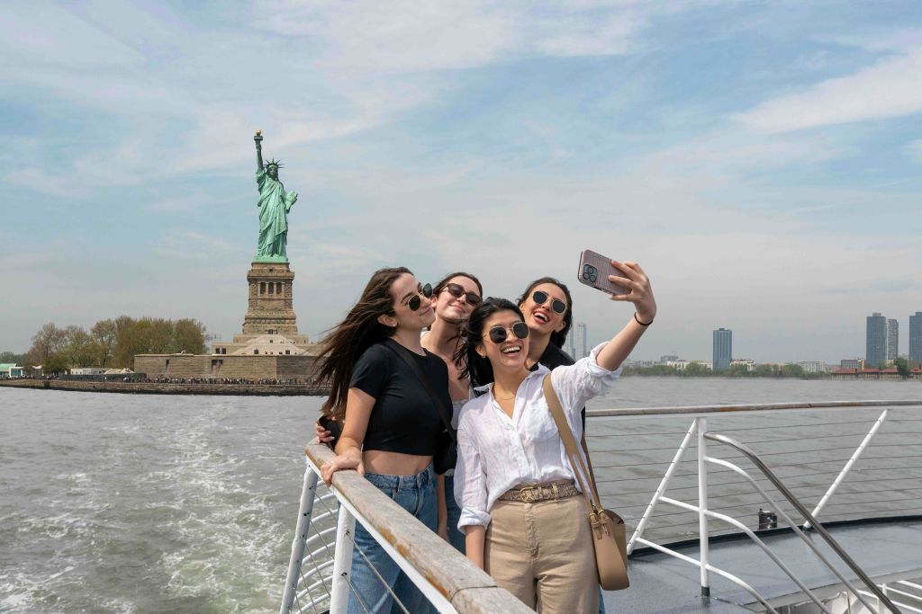 화창한 날, 네 명의 친구들이 자유의 여신상 근처에서 보트를 타고 셀카를 찍고 있습니다.