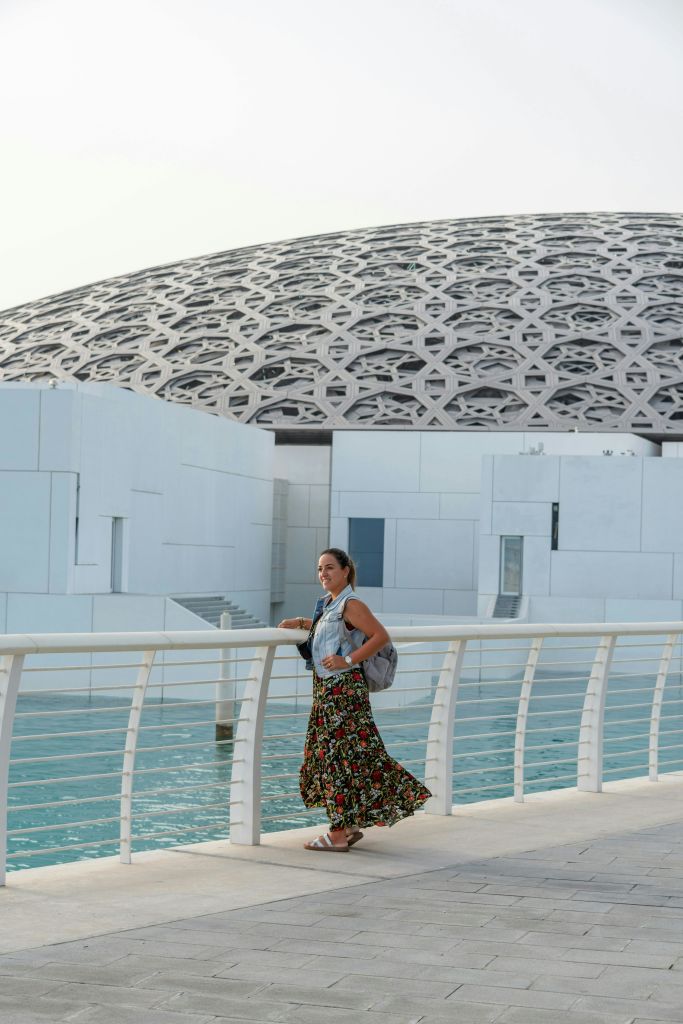 꽃무늬 치마를 입은 여성이 물과 기하학적 패턴의 돔을 배경으로 하얀 난간 근처에 서 있습니다.