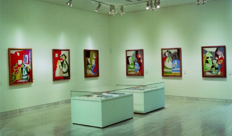 벽에 액자에 담긴 추상화와 중앙에 스포트라이트로 비춰진 두 개의 유리 디스플레이 케이스가 있는 아트 갤러리입니다.